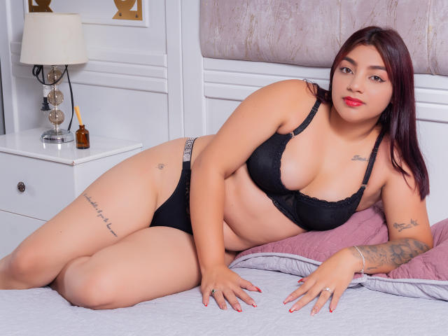 SamanthaSimon, with dark red hair, lying on a bed in black lingerie, showcasing her tits and ass, tattoos on her arm and thigh, red lipstick, white background.