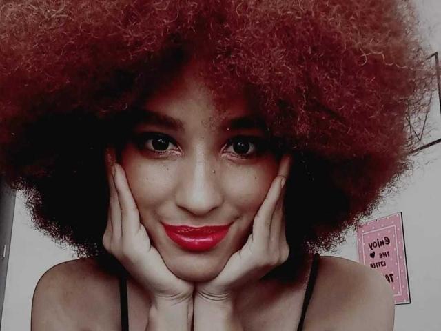 Violetttaa with curly red afro, hands on cheeks, smiling with red lipstick, brown eyes, and dark skin, wearing black straps, looking cute and sexy.