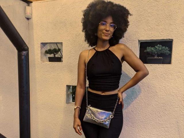 Violetttaa with dark afro, wearing glasses, black halter top, black pants, and silver chain purse, standing with hand on hip, smiling.