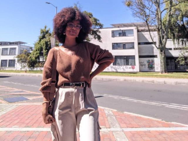 Violetttaa with big, fluffy afro, wearing glasses, brown ribbed sweater, and high-waisted beige pants, standing on brick sidewalk, hand on hip, sunny day, street in background.