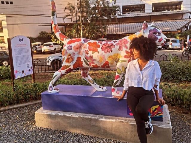 Violetttaa with curly brown hair, white shirt, black pants, and black shoes, sits next to a colorful floral-patterned horse statue on a purple platform.