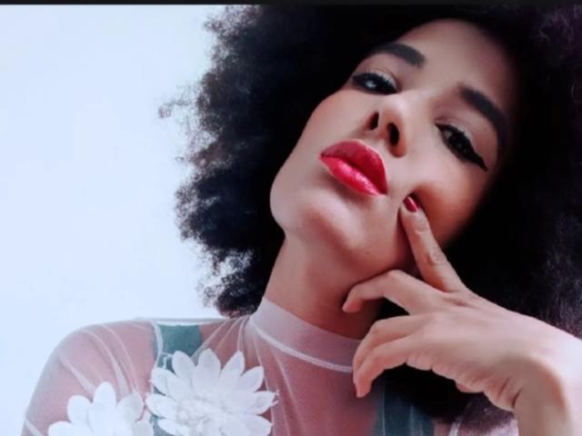 Violetttaa with curly black hair, red lipstick, and floral sheer top, touching her cheek with red-manicured fingers, looking down with a slightly pouty expression.