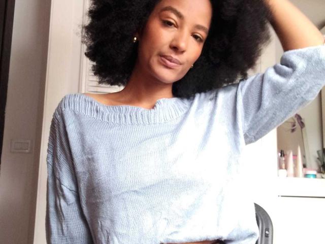 Violetttaa with curly black hair, wearing a light blue, off-shoulder sweater, hand in hair, subtle smile, small hoop earrings, soft makeup, standing in a bright room.