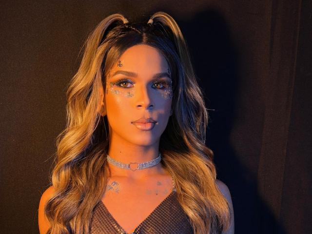 SammyLovely with long, wavy blonde hair, glittery face, blue eyes, and sparkly choker, wearing a mesh top, looking at camera with a slight smile.