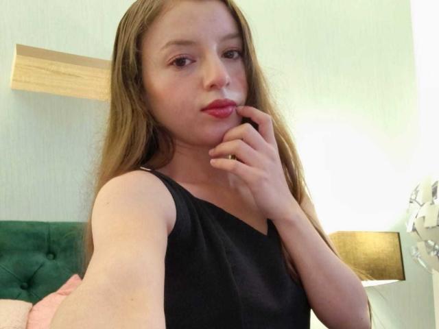 VioletaNova with long brown hair, black sleeveless top, pink lipstick, and finger touching her mouth, showing her bare shoulder and a gold ring on her finger.