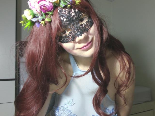 A photograph of AromaLove with long, red, wavy hair wearing a black lace mask and a floral headpiece with pink and white roses. She has a light skin tone and is smiling slightly. She wears a light blue, semi-transparent top with embroidered floral patterns. The lace mask has intricate floral patterns, and her hair is parted in the middle, falling over her shoulders. The lighting is natural, highlighting her features and the texture of her hair and mask.