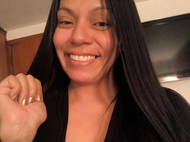 NataliaMargarita with long black hair, smiling, showing white teeth, wearing a black top, holding her fist up, nails painted silver, brown skin, brown eyes, in a selfie.