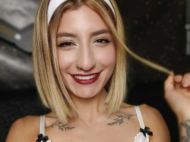 Kaiix with blonde bob, white headband, nose ring, red lipstick, smiling, holding hair, white bra with black bows, tattoos on chest. Boobs visible, tits small.