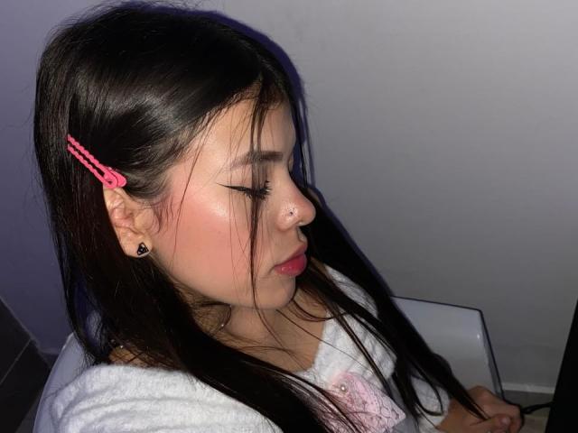 KeityRousse with long black hair, pink hair clips, nose ring, and white shirt, looking down with pink lips, and black ear studs, sitting in front of a laptop.