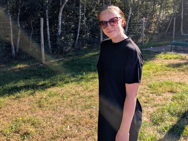 Larifari-hot with brown hair in a black dress and red sunglasses, standing in sunlit grass, smiling slightly, showing a slim, cute ass. Bright daylight, trees in background.