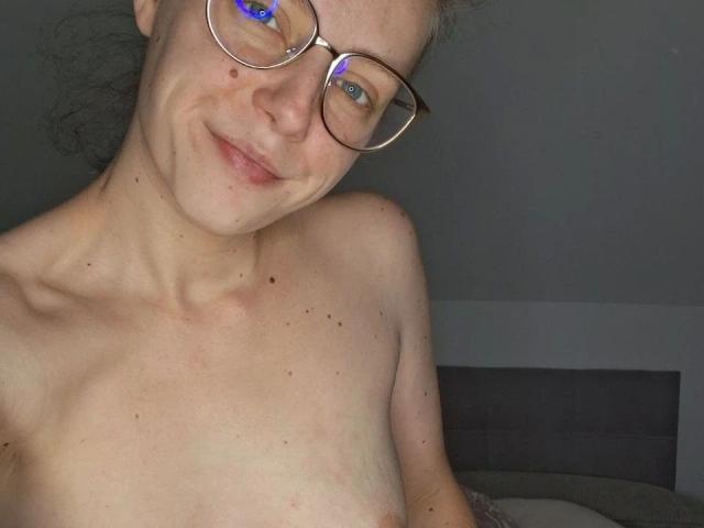 Larifari-hot with glasses, smiling at the camera, topless, showing small tits with a few moles, and blue eyes, in a dimly lit room.