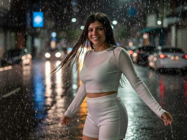 RilleyFoyx with wet, long brown hair, smiling, wearing a white long-sleeve crop top and high-waisted shorts, standing in the rain on a wet, illuminated street.