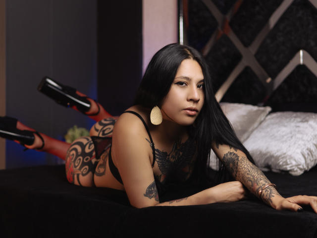 MaddieHandel with long black hair, tattoos on arms and legs, wearing black lingerie and high heels, lying on a bed, legs up, looking at camera, gold earrings.