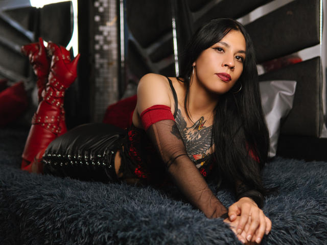 MaddieHandel laying on a blue furry bed, wearing a red off-shoulder top, black leather pants, and red boots, with tattoos on her chest and long black hair.