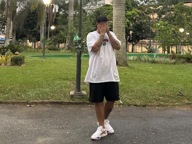 BigJota, wearing a white shirt and black shorts, stands on a sidewalk with hands covering mouth, looking confused. He has a medium build and is wearing white sneakers.