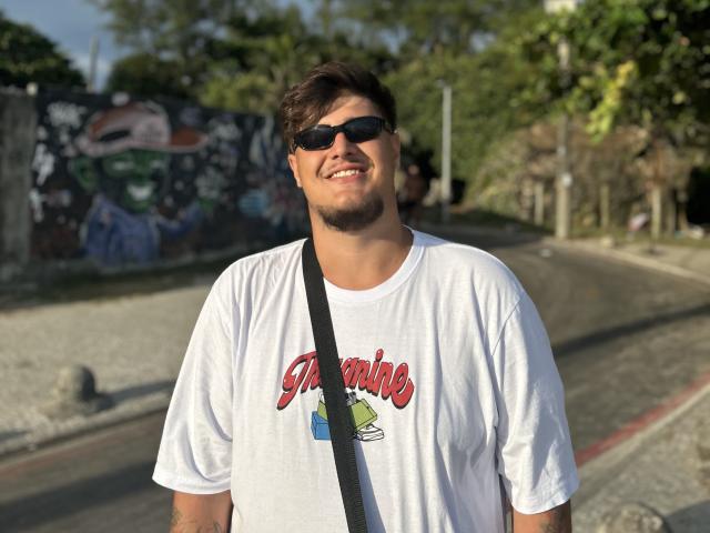 BigJota, chubby with dark hair and beard, wearing black sunglasses, white "Greenies" t-shirt, black strap, smiling on sunlit road, graffiti wall in background.