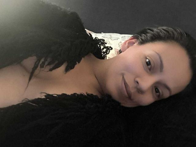 NataliaMargarita with black curly hair, lying on a black fluffy blanket, smiling at the camera, wearing a black feathered garment, exposing her tits. Soft lighting, calm expression.