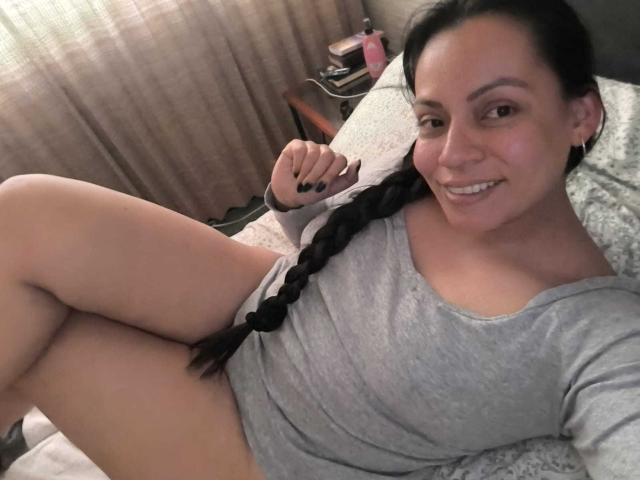 NataliaMargarita with long black braid, grey shirt, and white panties, smiling at camera, legs up, showing her pussy, on a bed, with black nail polish.
