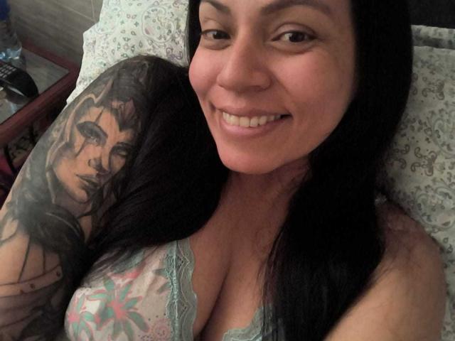 NataliaMargarita, smiling, with long black hair, tattooed arm, and large boobs, wearing lace lingerie, lying in bed, showing her cleavage and tattoo of a woman on her arm.