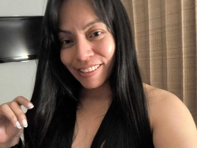 NataliaMargarita with long black hair, smiling, showing white nails, holding her hair, brown eyes, light brown skin, no clothes visible, in a selfie, looking cute.