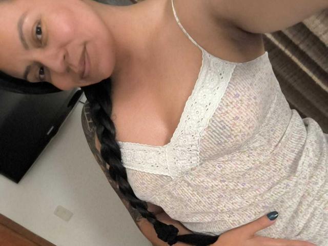NataliaMargarita with black braid, wearing white, see-through, lace-patterned dress, showing small tits, light skin, smiling, right arm raised, left hand on hip, blue nail polish.