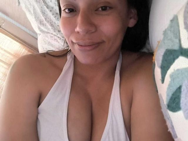 NataliaMargarita with a smile, wearing a white halter top, showing her tits, lying on a bed with green and white patterned pillows, brown hair, and light skin.