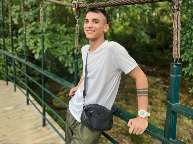 FaonLust, young dude with short brown hair, white tee, green pants, black bag, watch, and arm tattoo, leaning on rope bridge, smiling. Forest background.