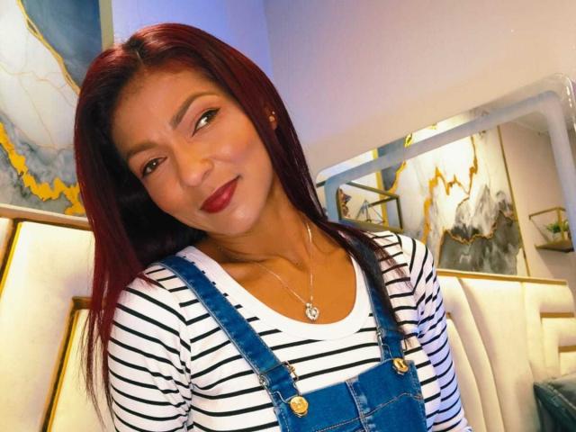 HannaPretty with red hair, wearing striped shirt and blue denim overalls, smiling, dark red lipstick, small necklace, sitting in a softly lit room.