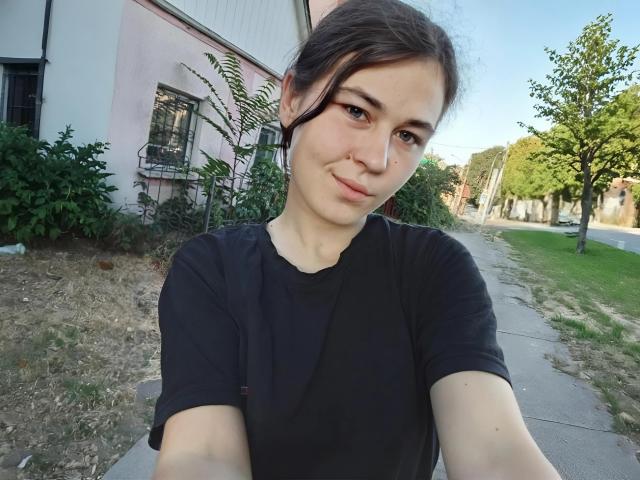 ToryTayler with a cute smile, brown eyes, black shirt, and black hair in a loose bun, taking a selfie outdoors on a sunny day. Her tits are small.