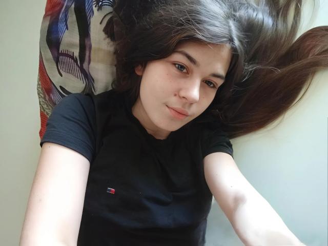 ToryTayler with long brown hair lying down, wearing a black shirt, looking at the camera with a slight smile, soft light on her face, slightly freckled skin.