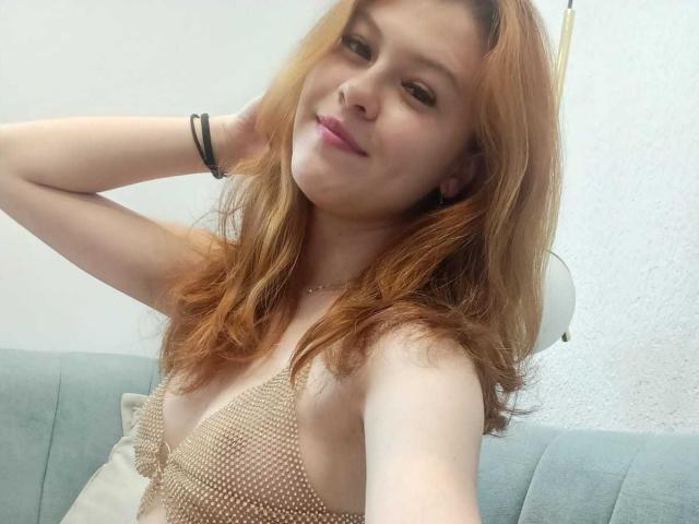 TaylorCarpert with red hair, smiling, wearing a beige mesh top that reveals her small tits. She's sitting on a blue couch, hand in hair, black bracelet on wrist.