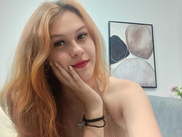 TaylorCarpert with orange hair, fair skin, and red lips, resting her hand on her cheek, wearing a halter top, necklace, and butterfly bracelet, smiling softly.