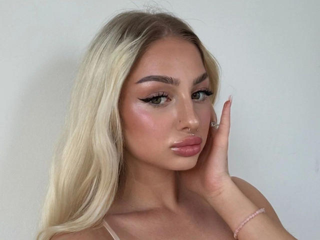 StellaLiebe-hot with blonde hair, green eyes, and pink lips, touching her cheek with a hand. She has a nose ring and pink bracelet, looking slightly to the side.