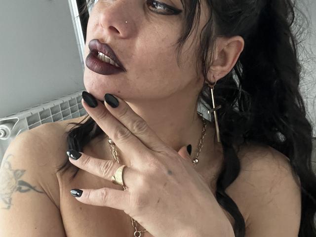 Cyberxxx with dark lipstick, black nails, gold hoop earrings, ring, and chain, touching her neck, showing tattoos, and a nose piercing, with long black hair.