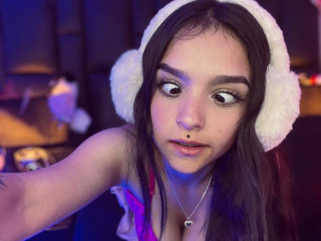 MarietteJones with wide eyes, black eyeliner, and a nose piercing, wearing a fluffy white hat, and a necklace with a heart pendant, has her tits slightly visible.