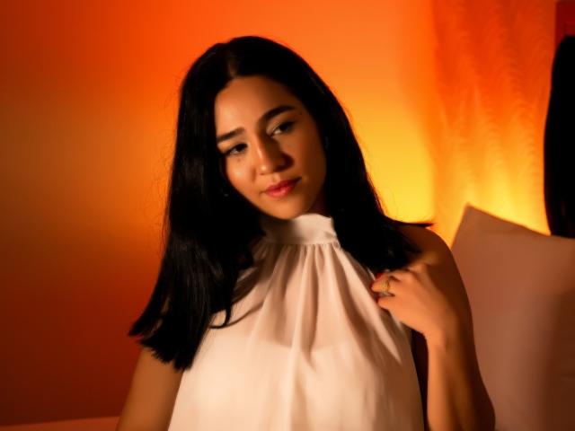 SamantaFire with black hair, wearing a white sleeveless top, holding her shoulder, orange light in the background, subtle smile, soft light on her face.
