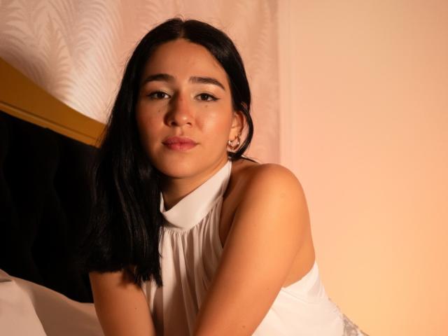 SamantaFire with black hair, brown eyes, and light brown skin, wearing a white halter top, looking at the camera with a slight smile, her tits partially visible.