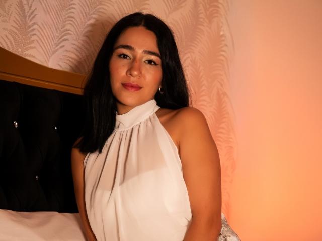 SamantaFire with medium brown skin, black hair, and pink lipstick, wearing a white halter top, sitting on a bed, smiling softly. Her small tits are slightly visible.