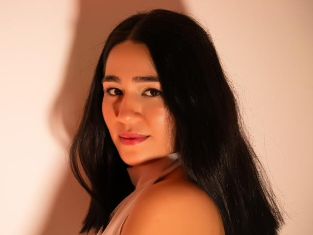 SamantaFire with long black hair, light brown skin, and red lipstick, looking over her shoulder, wearing a white halter top, showing her bare shoulders and subtle smile.