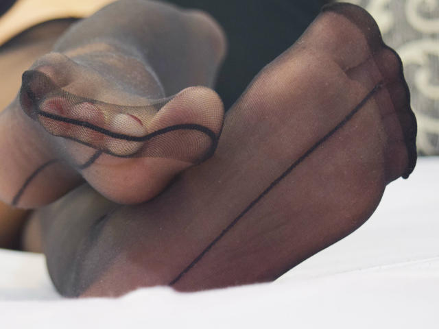 Les pieds de SelinaKat, portant des bas noirs transparents, laissant entrevoir ses orteils et la couture du tissu, sont en l'air, avec un orteil légèrement recourbé sur le côté droit..
