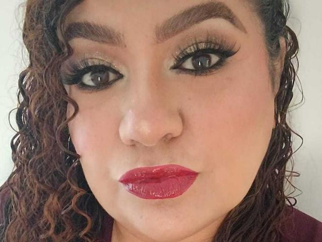 SeleneFhox69's face with curly brown hair, thick eyebrows, dark eyeliner, long lashes, and shiny red lips. She's looking directly at the camera with a slight pout.