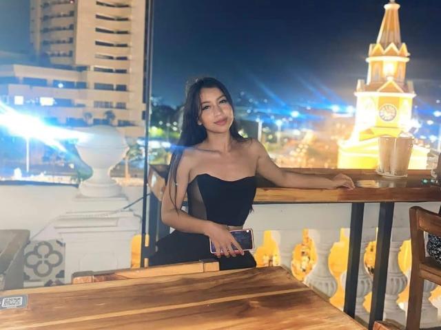 SaraColling with long black hair, wearing a strapless black dress, sitting at a rooftop table, holding a phone, smiling, night city lights in the background.