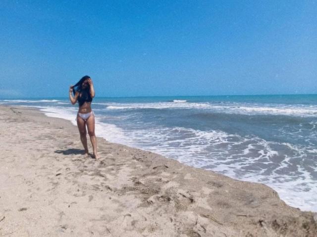 SaraColling stands on a beach, wearing a white bikini, her long dark hair covering one eye, looking at the ocean with gentle waves. Her ass is prominently visible.