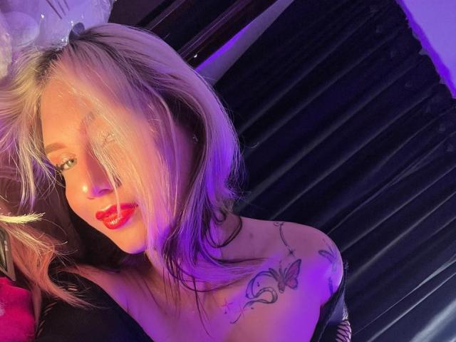 MariyLu with blonde hair, red lipstick, tattoo on right shoulder, black off-shoulder top, blue-purple lighting, slightly messy hair, smiling, looking at camera, soft light, casual setting.