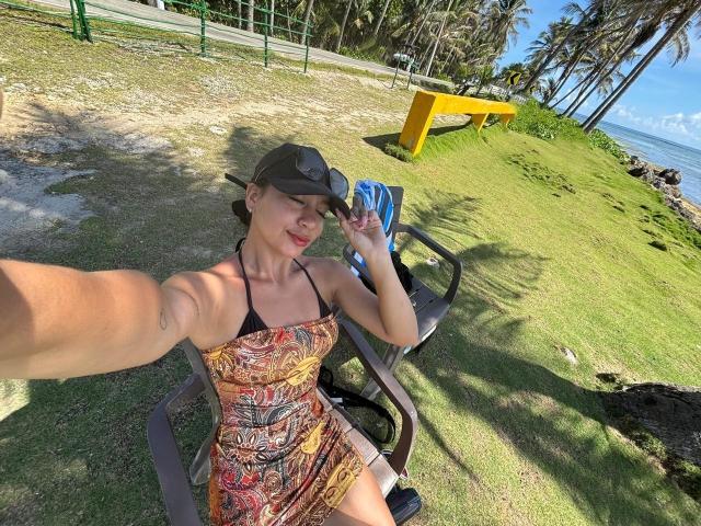 SaraColling in a patterned dress, tan, and small tits, sitting on a chair, wearing a cap, holding a phone, taking a selfie, with a green grassy hill.