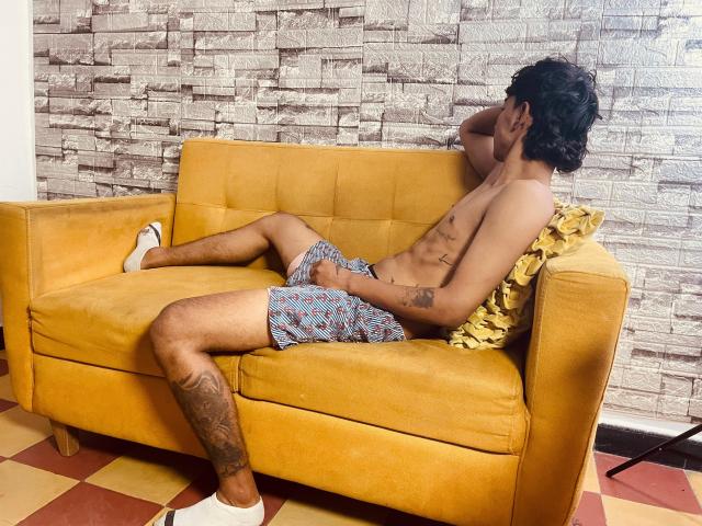 Shirtless Bracth with tattoos, wearing patterned shorts and white socks, lounges on a yellow couch, legs spread, hand on crotch, yellow pillow, stone wall background, checkered floor.