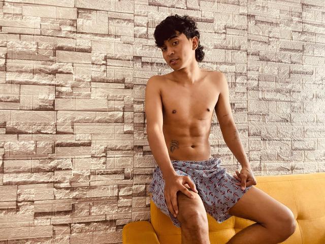 Shirtless Bracth with a small tattoo on his side, wearing blue and red patterned shorts, sitting on a yellow couch, hands on knees, against a textured stone wall.