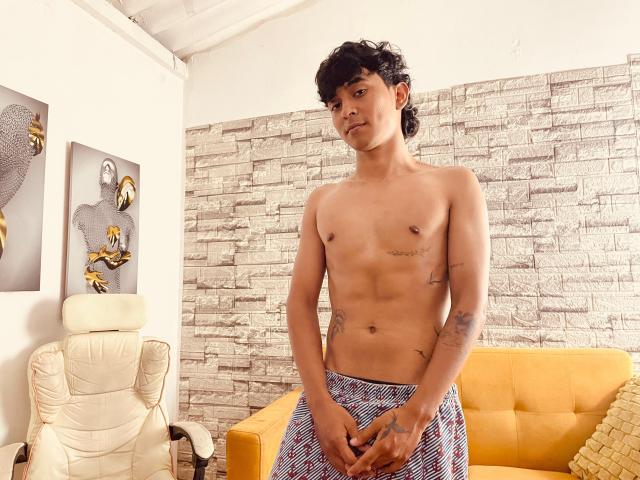 Shirtless Bracth stands in a room with a textured white brick wall. Bracth has a slender, toned build, small nipples, and dark curly hair. Bracth wears blue and white checkered pants, hands are relaxed in front. Tattoos are visible on Bracth's right arm and left side. Yellow couch and white chair are in the background. Two bird-themed paintings hang on the left. The room has a white ceiling. Warm, natural light.