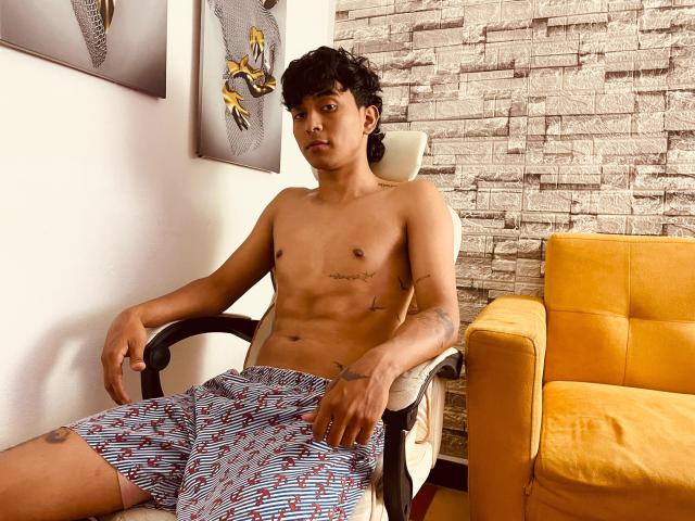 Bracth is a shirtless, young Asian man with curly black hair, sitting in a black chair. He has a lean, muscular build with visible abs and tattoos on his arms and left thigh. He's wearing blue and white striped shorts with red dots. The yellow armchair to his right contrasts with the textured, white stone wall behind him. Two abstract paintings hang on the wall to his left. The image is well-lit, emphasizing Bracth's relaxed, slightly bored expression.