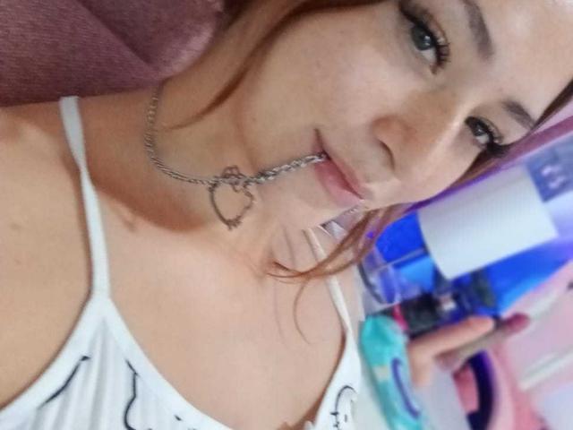 SamanthaCollen with pink hair, wearing a white tank top, has a metal chain with a heart-shaped ring in her mouth, showing her tits. Her eyes are slightly open.